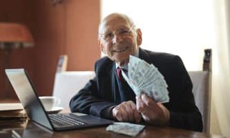 Happy senior businessman holding money in hand while working on laptop at table