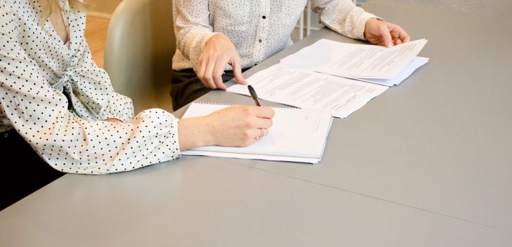 woman signing on white printer paper beside woman about to touch the documents